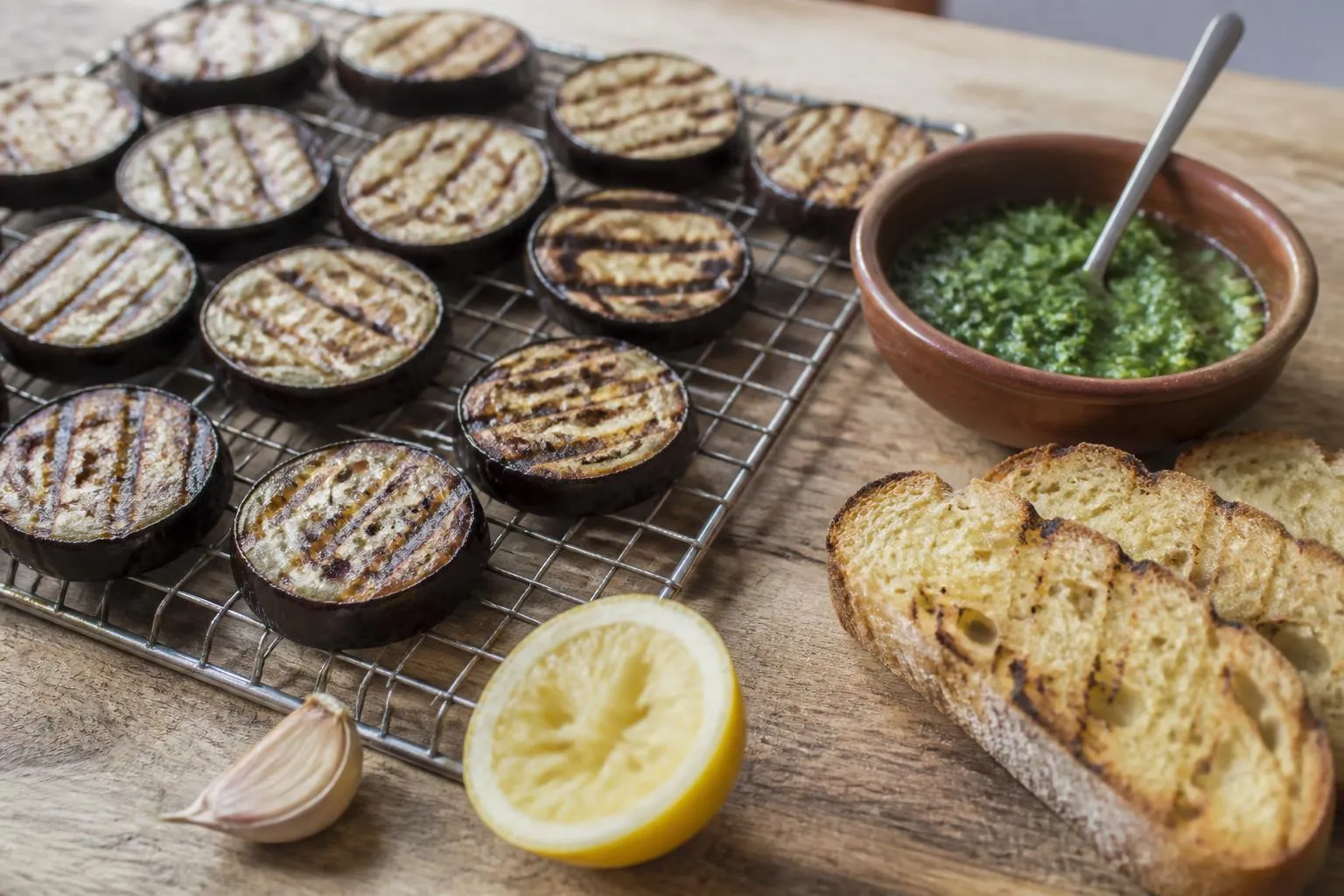 Rondelles d'aubergine marquées au gril posées sur une grille, à côté d'un bol de persillade et de tranches de pain de campagne grillées.