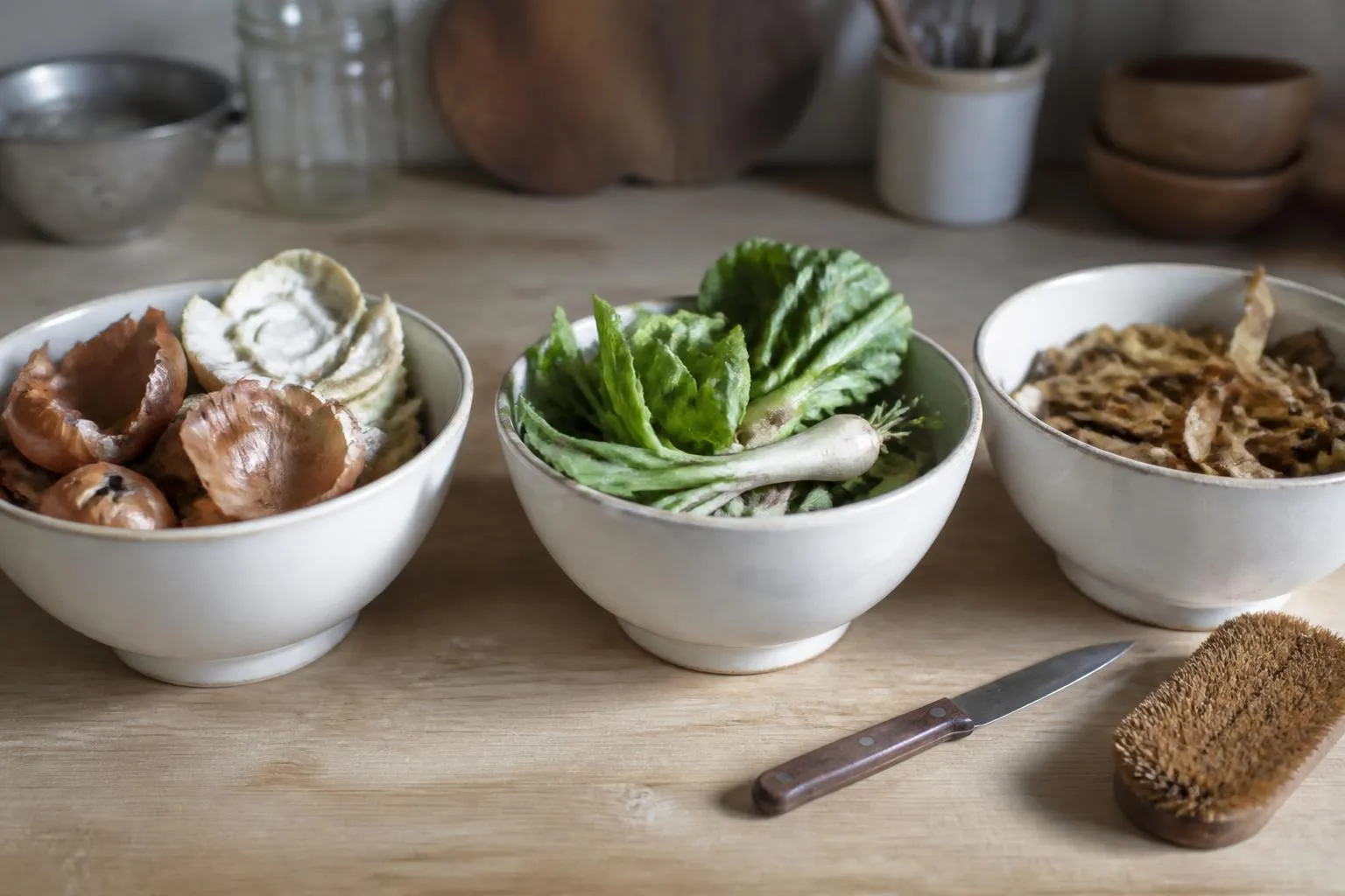 Trois saladiers alignés sur une table de cuisine, remplis de fanes, de pelures et de trognons de légumes