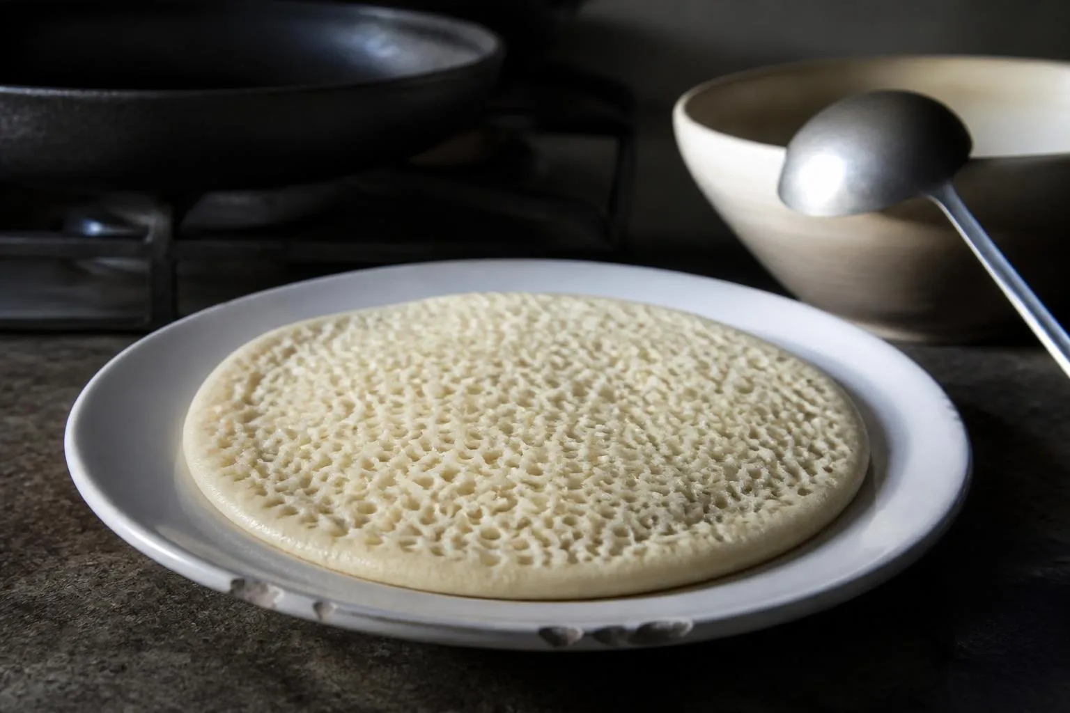 Un baghrir posé sur une assiette blanche, sa face supérieure criblée de cavités ouvertes, à côté d'une poêle noire et d'une louche.