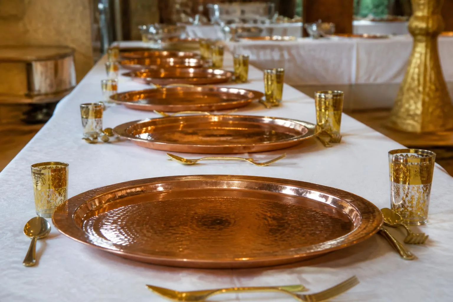 Une longue table de banquet dressée avant le service, alignement de grands plats ronds vides, de verres à thé et de couverts, sous une nappe blanche.